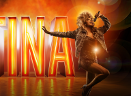 Tina Turner står foran et stort lysende skilt, hvor der står TINA på. Hun har en mikrofon i hånden.